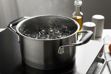 Cooking pot with boiling water and stove on light table against grey background, closeup
