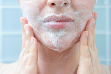 Fototapeta premium Young woman applying cleansing foam to her face close-up against a background of blue tiles.