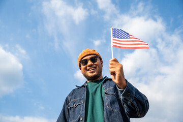 Portrait of cheerful young man looking forward while standing proudly with holding USA flag over his head against wind and blue sky, happy american holding the United states flag celebration
