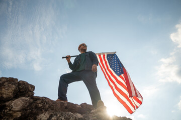Portrait of cheerful young man looking forward while standing proudly with holding USA flag over his head against wind and blue sky, happy american holding the United states flag celebration