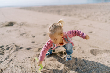 Little girl squats on the beach and digs the sand with a toy shovel with her hand out to the side