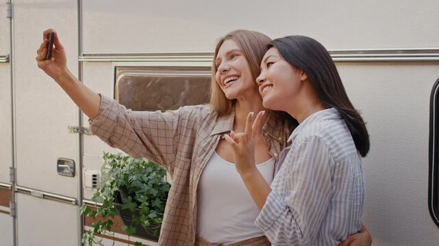 Diverse girlfriends gather outdoors during summer for a weekend camping trip. They share laughter and camaraderie while capturing joyful moments on their phones.