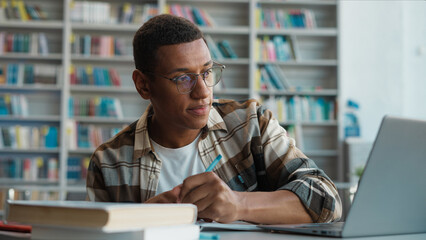 Fototapeta premium African American man working studying laptop desktop computer e-learning education male student copying writing notebook looking monitor preparing exam guy library technology knowledge college diploma