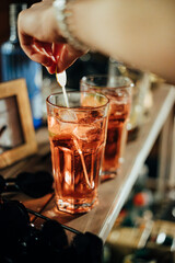 A close-up of a hand stirring an iced red cocktail in a tall glass, surrounded by drinks and bar elements, capturing a lively celebratory vibe