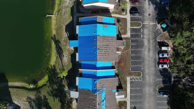 Townhouses or row of homes with blue foil on roof In town of Florida. Hurricane destroying rooftop of buildings. Aerial top down shot. Lake in garden and parking cars. Suburb district of Bradenton.