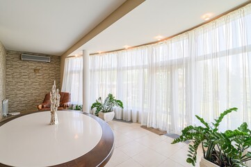 bright, stylish living room with a round table, a leather armchair, and large windows draped with sheer white curtains. The room is decorated with plants and features textured walls