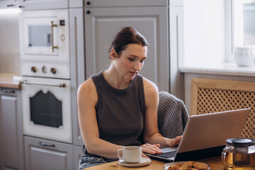 A beautiful brunette woman is in the kitchen at the table working on a laptop