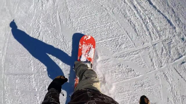 First-person view of snowboarding on a groomed slope. Red snowboard, long shadow, sunny day. Winter sports action, snow texture, close-up shot.