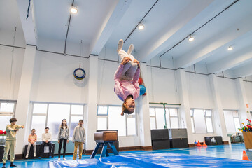 Teenage girl practicing backflip over vaulting horse in school gymnastic class, with classmates...