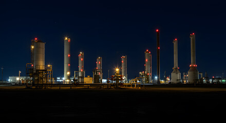 Illuminated industrial complex against a dark night sky, towers and pipes with glowing red lights.