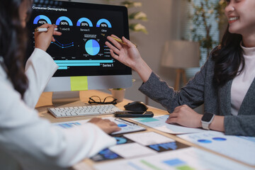 Two businesswomen are collaborating on consumer demographics, analyzing data displayed on a...