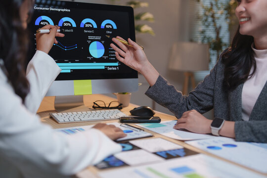 Two businesswomen are collaborating on consumer demographics, analyzing data displayed on a computer screen and using printed reports to gain insights into market trends and customer behavior