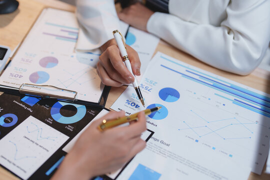 Two businesswomen are collaborating on analyzing financial charts and statistics, using pens to point at key data points and discuss performance metrics