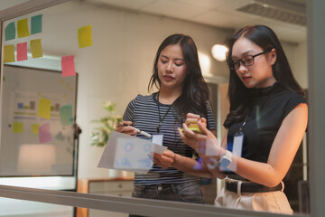Two young businesswomen are using sticky notes on a glass wall and discussing work while reviewing documents and using mobile phone in a modern office