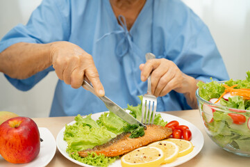 Asian senior woman patient eating Salmon steak breakfast with vegetable healthy food while sitting and hungry on bed in hospital.