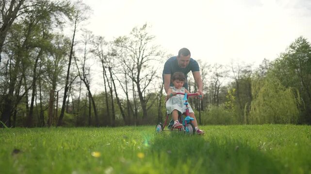 Father helps toddler ride bike park. Family teaching toddler to ride bike outdoors. Kid learning to ride bike with father. Joyful father teaches toddler bike in family park. Kid father family outdoors