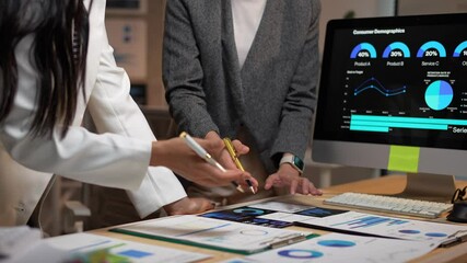 Two businesswomen collaborate, analyzing consumer demographics displayed on a computer screen and printed charts, pointing and discussing data insights to optimize marketing strategies
