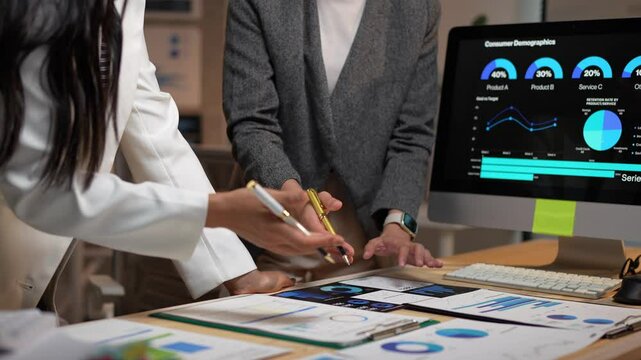 Two businesswomen collaborate, analyzing consumer demographics displayed on a computer screen and printed charts, pointing and discussing data insights to optimize marketing strategies