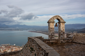A stone arch with a hanging bell overlooks a panoramic view of the bay and distant mountains under a cloudy sky. Captured from the historic Palamidi Fortress in Nafplio, Greece, this image blends anci