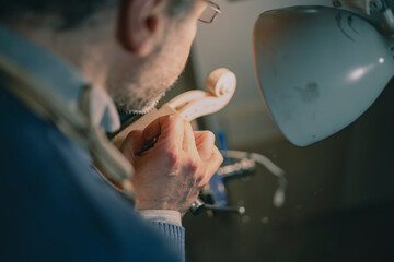 Inside a Cremonese workshop, a skilled luthier chisels a violin scroll held in a blue vise under soft lighting, wearing a leather apron, working with deep concentration and artisan mastery.