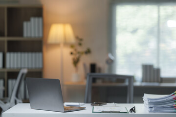 Laptop, documents, clipboard, eyeglasses, and smartphone arranged on a white desk in a cozy, modern home office, with a blurred background of a bookcase and window