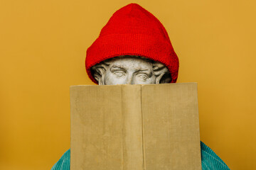 Antique statue of a young man in a red hipster hat hiding behind a book