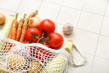 Mesh bag with different fresh vegetables on light tile background, closeup