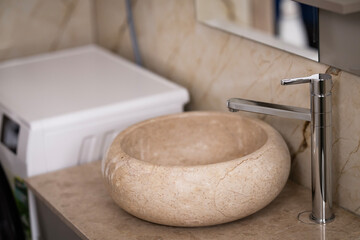 Close-up of a cracked marble bathroom sink with a modern chrome faucet. Visible damage on the stone basin, located next to a washing machine in a tiled interior.