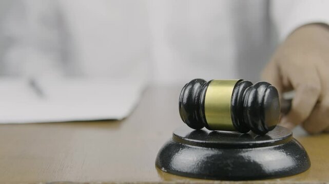 A gavel in the foreground, with a person in white clothing signing a document in the background, then striking the gavel with their left hand, symbolizing the finalization of a legal decision or agree