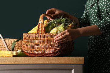 Woman holding wicker basket with fresh corn cobs on wooden table against green background