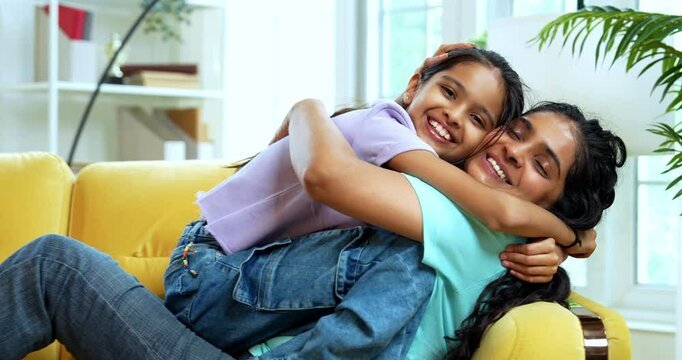 Indian Asian young mother joyfully playing seesaw with her little daughter on sofa in stylish modern living room, enjoying fun bonding moments, happiness, laughter, and indoor quality time together