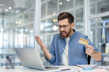 A frustrated businessman in a modern office, holding a credit card and staring at his laptop with...