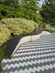A bench is on a brick walkway with a tree behind it. The bench is black and the walkway is made of bricks