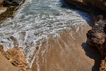 Foam traces spread across wet sand as waves reach a small cove surrounded by rugged rocks with mossy patches on the left side; tourism;