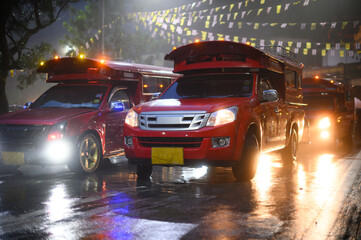 Rod Dang or red car waits for passengers on a rainy street in front of Wat Phra That Doi Suthep in Chiang Mai. The reflection of lights glistens on the wet road during the rainy season.
