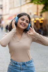 Happy Indian young woman in wireless headphones dancing listening favorite energetic disco music in smartphone dancing outdoors. Hispanic girl walking on urban city street. Town lifestyles. Vertical