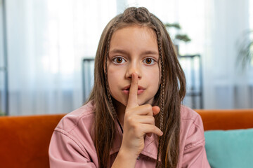 Fototapeta premium Shh be quiet please. Portrait of preteen girl child presses index finger to lips makes silence hush gesture sign do not tells gossip secret. Little cute teenage female sitting on sofa in living room.