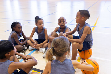 Naklejka premium In school gym, multiracial children sitting on floor listening to teammate, holding basketball