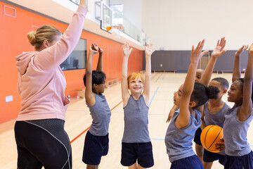 In school gym, diverse children raising hands with female teacher, holding basketball, smiling