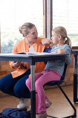 Female teacher helping young girl with schoolwork in classroom, engaging in discussion