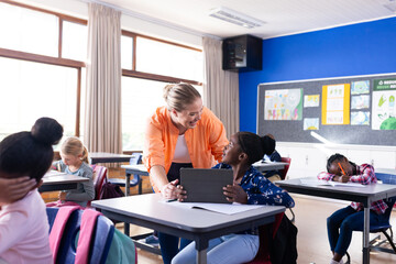 Female teacher helping african american girl with tablet in classroom, other students in school