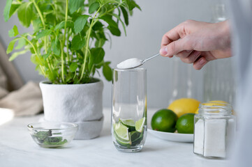 Making homemade summer drink, person pouring sugar in glass with lime and mint, mojito making process at home
