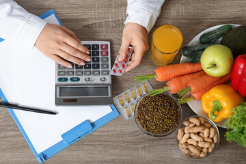 Young female nutritionist with vitamins, calculator and healthy food writing diet plan in office,...