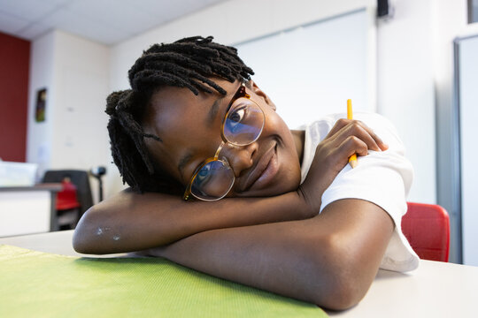In school, smiling african american boy with glasses holding pencil, resting on desk