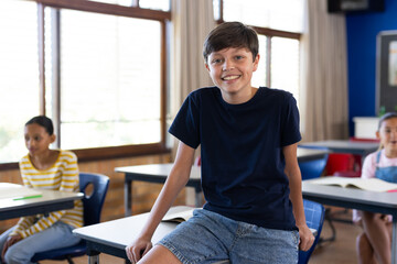 Smiling boy sitting on desk in classroom with students in background at school