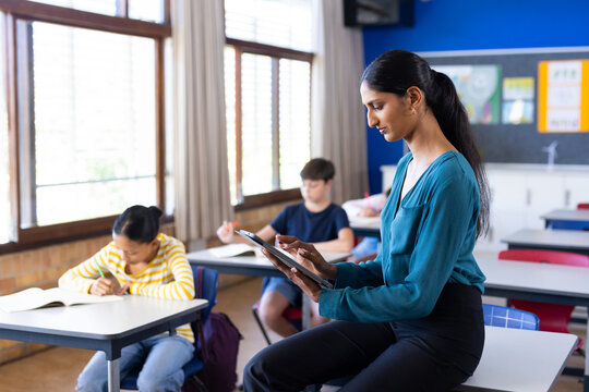 In school, Indian female teacher using tablet while students studying in classroom, copy space