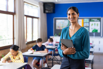 In school classroom, holding tablet, smiling indian female teacher with students, copy space