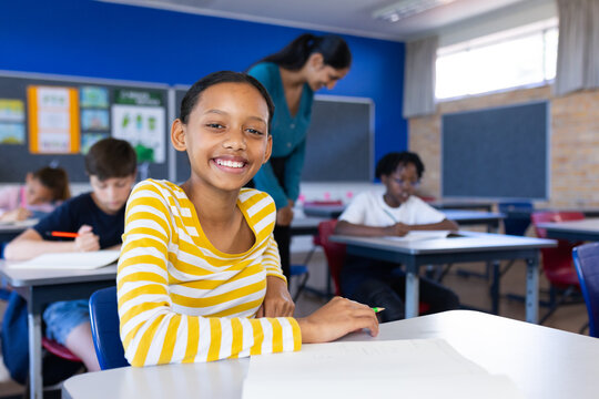 In school, smiling girl sitting at desk with classmates and female teacher in classroom, copy space