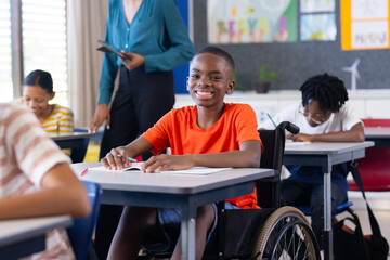 In school, boy in wheelchair smiling and writing at desk in classroom