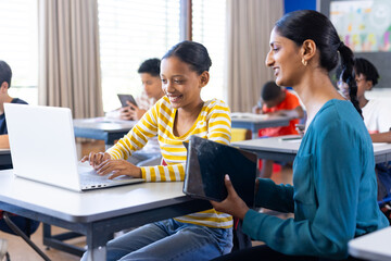 In school, Indian female teacher helping student using laptop in classroom, smiling together